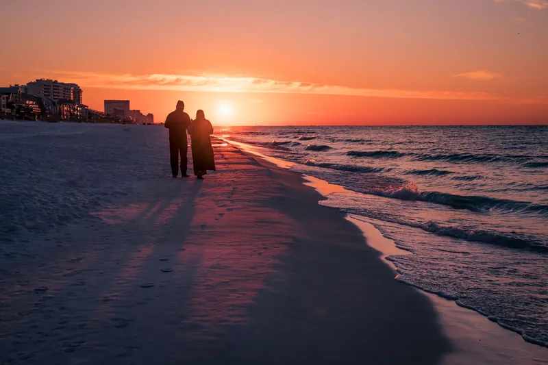 Two people walking along a wide beach at sunset, seen as silhouettes