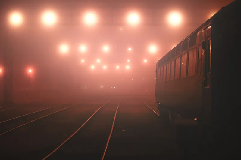 Railway tracks in thick fog at night under rows of glowing lamps, a train on the right
