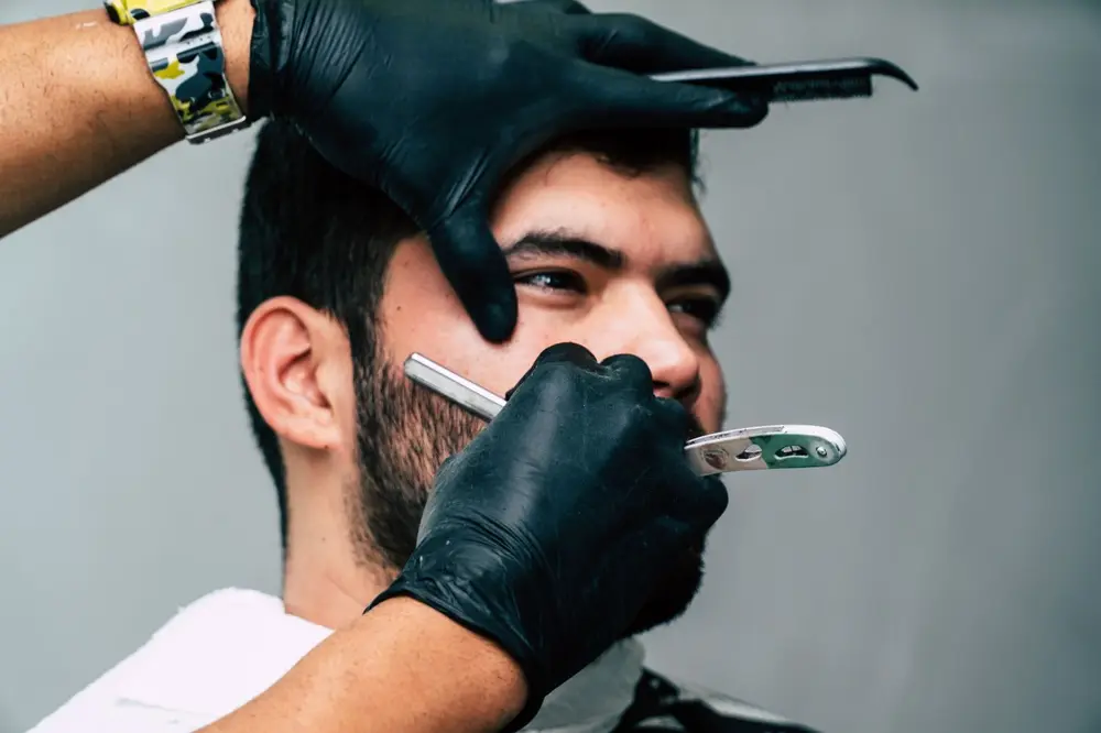A barber in black gloves shaving a client's cheek with a straight razor