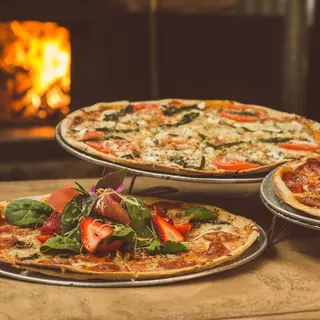 Pizzas with toppings on a table in front of a wood fire