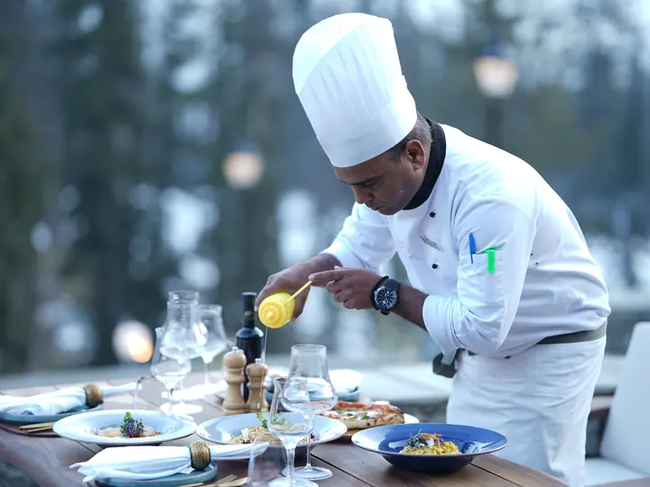 A chef in a white hat plating a dish at an outdoor table