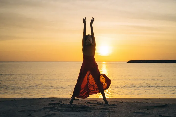 Silhouette of a model in a red dress with arms raised on a beach at sunset