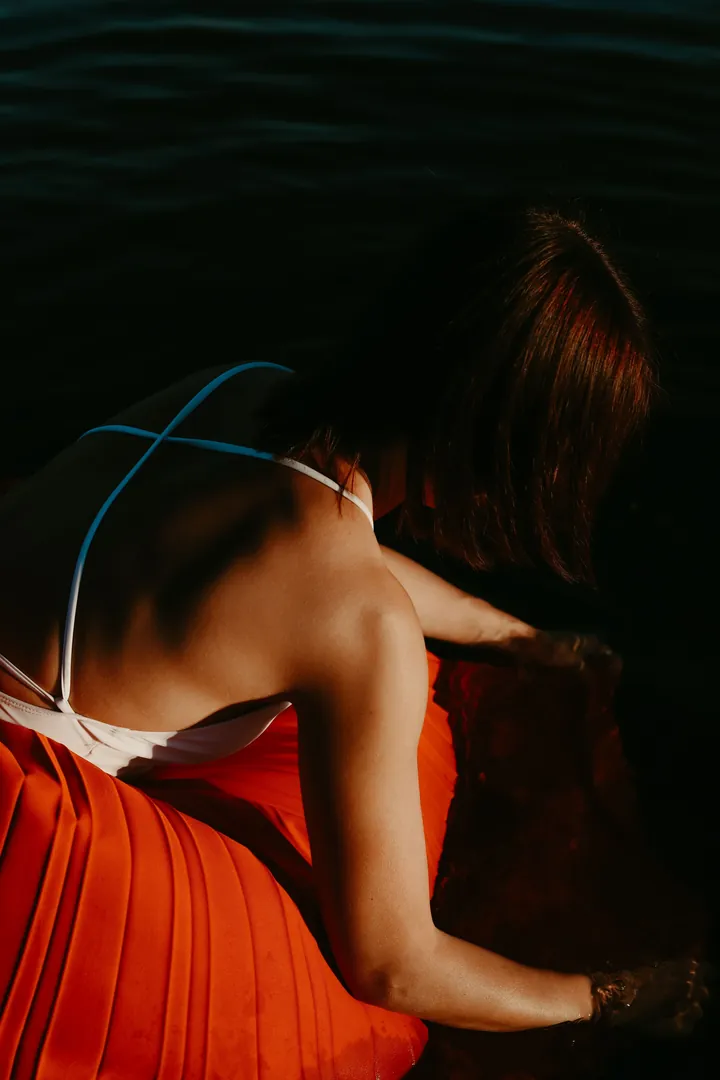 Model in a pleated vermilion skirt turning away from the camera, dark water behind her