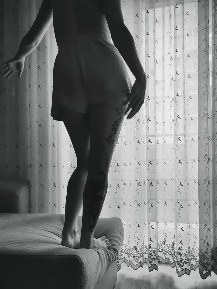 Black and white photo of a model standing by a lace curtain in window light